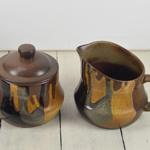 May include: A brown and tan ceramic creamer and sugar bowl set with a drip glaze design. The creamer has a handle and the sugar bowl has a lid.