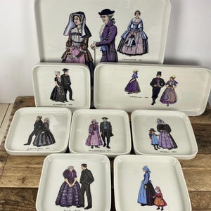 May include: A collection of white ceramic trays and plates featuring illustrations of people in traditional European clothing. The illustrations are in color and include text. The collection includes a large rectangular tray, a smaller rectangular tray, and several square plates.