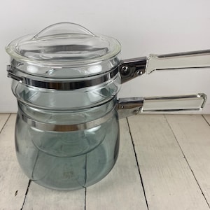 May include: A clear glass double boiler with a matching lid. The pot has two handles with clear plastic grips and is held together with silver metal bands. The glass has a slight blue tint. The double boiler is designed for cooking.