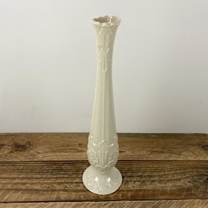 May include: A tall, cream-colored ceramic vase with a fluted, slender body and a flared, scalloped rim. The vase features ornate, raised leaf and scroll detailing around the base and midsection. It stands on a round, pedestal-style base.