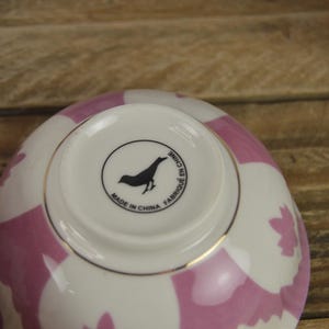 May include: A white ceramic bowl with a pink and white geometric pattern. The bowl has a gold rim and a black bird logo on the bottom that reads "Made in China Fabriqu&eacute; en Chine".