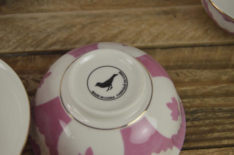 May include: A white ceramic bowl with a pink and white geometric pattern. The bowl has a gold rim and a black bird logo on the bottom that reads "Made in China Fabriqu&eacute; en Chine".