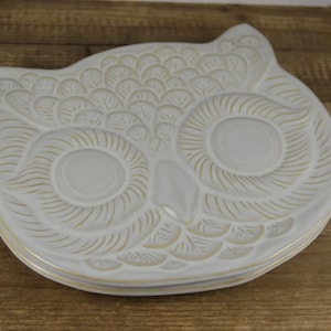 May include: White ceramic owl-shaped serving dish with gold-colored details. The owl's eyes and feathers are outlined in gold.