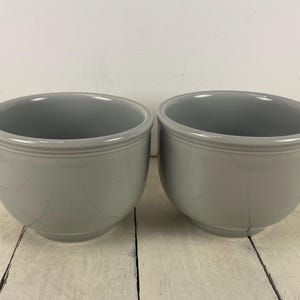 May include: Two gray ceramic bowls with a smooth, glossy finish. Each bowl has a rounded shape with a slightly flared rim and a decorative band near the top. The bowls are set against a white wooden surface.