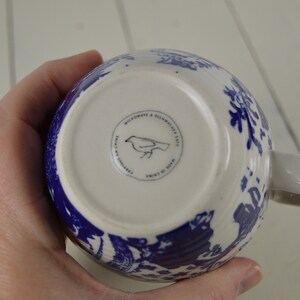 May include: White ceramic mug with a blue floral pattern. The bottom of the mug features a circular stamp with a bird illustration and the text "MICROWAVE & DISHWASHER SAFE". The mug has a white handle.