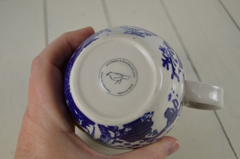 May include: White ceramic mug with a blue floral pattern. The bottom of the mug features a circular stamp with a bird illustration and the text "MICROWAVE & DISHWASHER SAFE". The mug has a white handle.