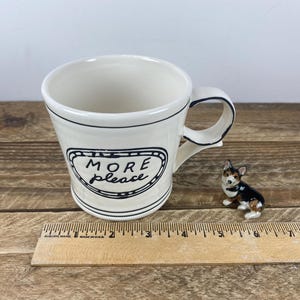 Puede incluir: Una taza de cer&aacute;mica color crema con borde y asa negros, con las palabras "MORE please" en un marco negro decorativo. Una peque&ntilde;a figura de perro de cer&aacute;mica est&aacute; al lado de la taza. Una regla de madera est&aacute; en primer plano.
