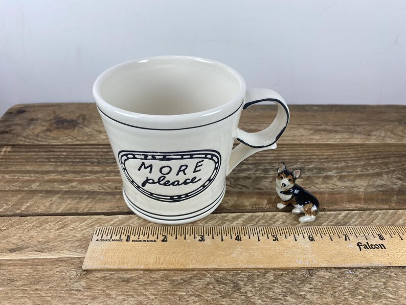 Puede incluir: Una taza de cer&aacute;mica color crema con borde y asa negros, con las palabras "MORE please" en un marco negro decorativo. Una peque&ntilde;a figura de perro de cer&aacute;mica est&aacute; al lado de la taza. Una regla de madera est&aacute; en primer plano.