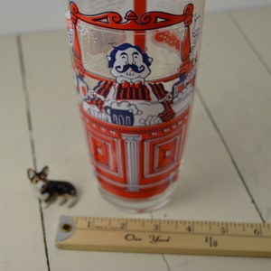 May include: A red and white glass cocktail shaker with a cartoon illustration of a bartender pouring a drink. The bartender is wearing a red and white striped shirt and a bow tie. The shaker is about 7 inches tall.