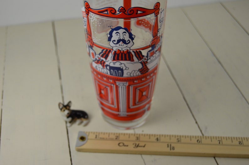 May include: A red and white glass cocktail shaker with a cartoon illustration of a bartender pouring a drink. The bartender is wearing a red and white striped shirt and a bow tie. The shaker is about 7 inches tall.