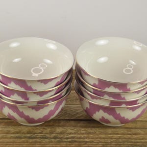 May include: Set of six pink and white ceramic bowls with a gold rim. The bowls are stacked in two piles of three. The bowls have a chevron pattern.