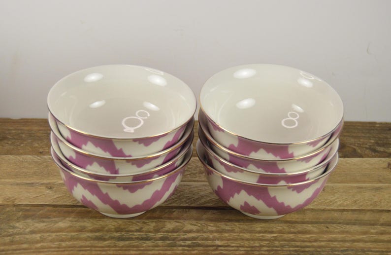 May include: Set of six pink and white ceramic bowls with a gold rim. The bowls are stacked in two piles of three. The bowls have a chevron pattern.