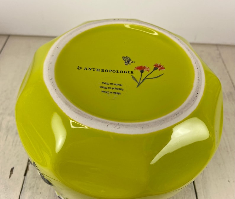 Może przedstawiać: Chartreuse ceramiczna miska z białym obramowaniem. Wnętrze miski zawiera napis "by ANTHROPOLOGIE" i mały kwiatowy wz&oacute;r. Sp&oacute;d miski jest oznaczony "Made in China" i "Hecho en China".
