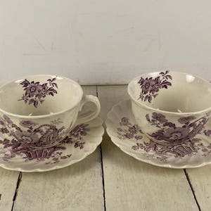 May include: Two matching teacups and saucers. The cups and saucers are white with a purple floral and basket design. The saucers have a scalloped edge. The cups have handles.