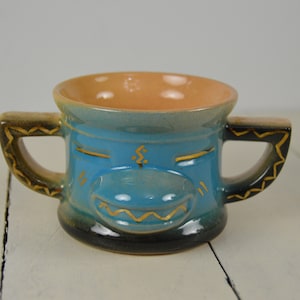 May include: A blue ceramic mug with two handles and a stylized face design. The face has gold accents and a closed-eye expression.