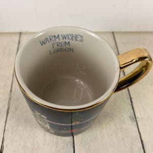K&ouml;nnte beinhalten: Eine Keramik-Tasse mit goldenem Henkel und Rand, mit einer dunkelblauen Au&szlig;enseite mit einem Londoner Stadtbild-Design. Der Innenraum ist cremefarben, mit dem Text "WARM WISHES FROM LONDON" in Blau.