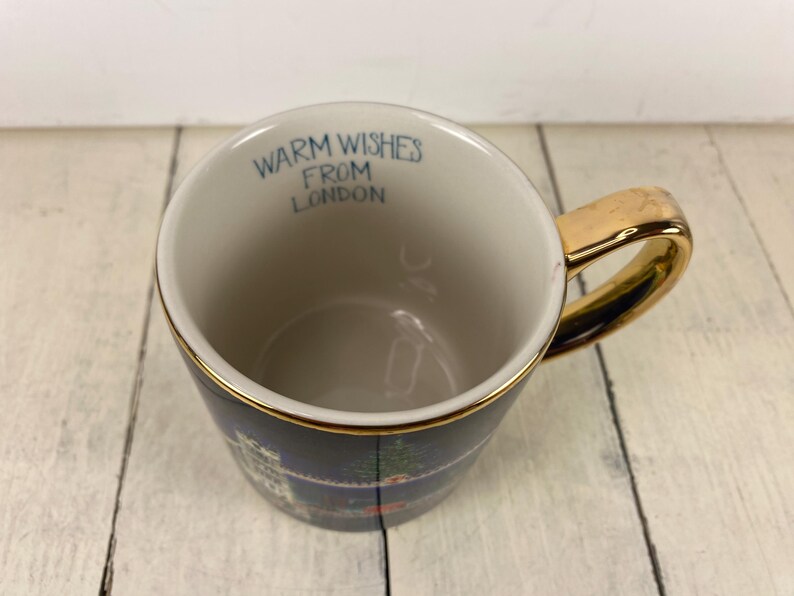 K&ouml;nnte beinhalten: Eine Keramik-Tasse mit goldenem Henkel und Rand, mit einer dunkelblauen Au&szlig;enseite mit einem Londoner Stadtbild-Design. Der Innenraum ist cremefarben, mit dem Text "WARM WISHES FROM LONDON" in Blau.