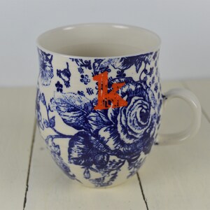 May include: White ceramic mug with a blue floral pattern and an orange letter "K". The mug features a handle and a detailed design of roses and other flowers. The mug is suitable for hot or cold beverages.