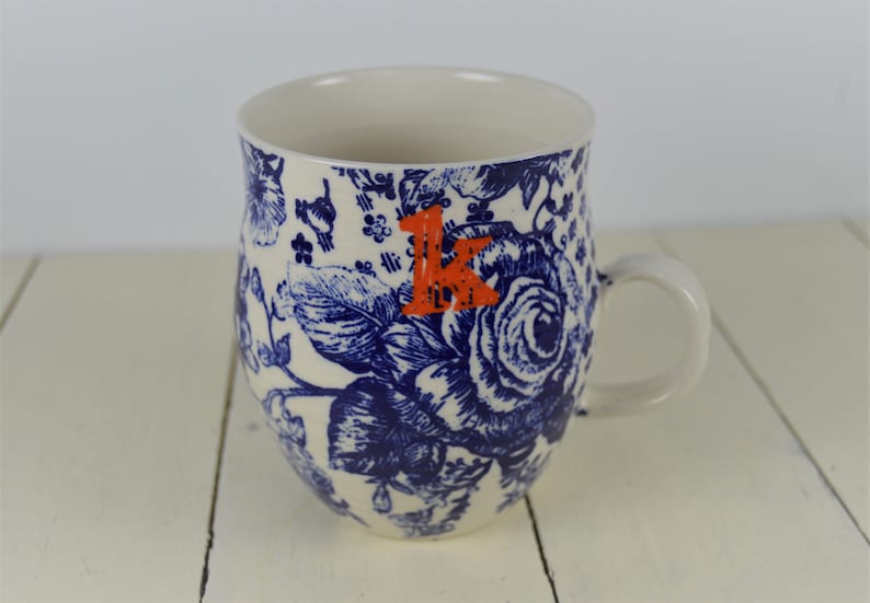 May include: White ceramic mug with a blue floral pattern and an orange letter "K". The mug features a handle and a detailed design of roses and other flowers. The mug is suitable for hot or cold beverages.