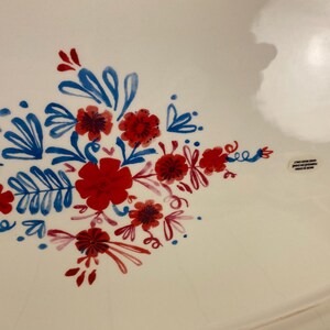 Op de afbeelding: Witte keramische serveerschaal met een handgeschilderd bloemmotief in rood en blauw. De bloemen en bladeren zijn gerangschikt in een decoratief patroon. De schaal heeft een licht gebogen rand.