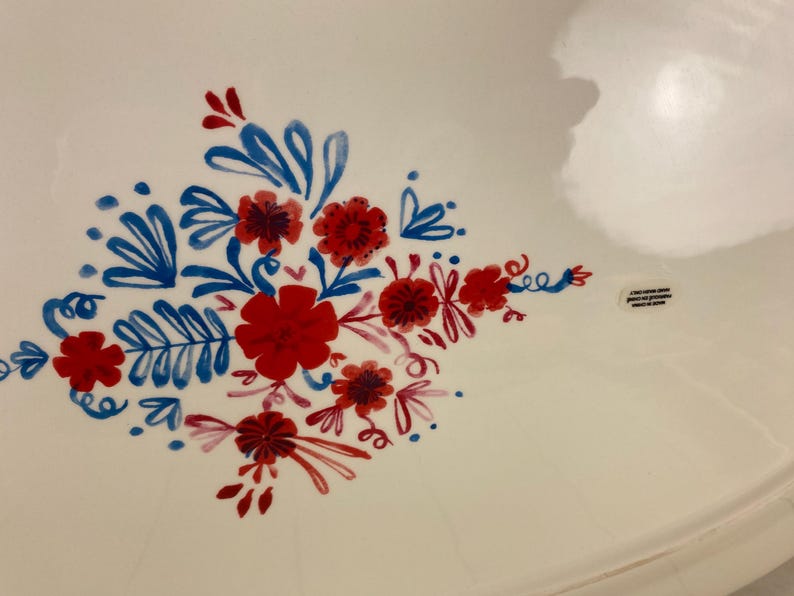 Op de afbeelding: Witte keramische serveerschaal met een handgeschilderd bloemmotief in rood en blauw. De bloemen en bladeren zijn gerangschikt in een decoratief patroon. De schaal heeft een licht gebogen rand.