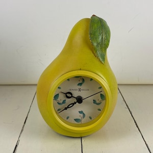 May include: A pear-shaped table clock with a yellow body and a green leaf at the top. The clock face is white with small green leaf illustrations, black hands, and the text Howard Miller printed in black at the center of the dial.