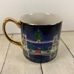 K&ouml;nnte beinhalten: Eine Keramik-Tasse mit goldenem Henkel und Rand. Die Tasse hat einen dunkelblauen Hintergrund mit einer Weihnachtsszene, darunter ein geschm&uuml;ckter Baum, Geb&auml;ude und eine Br&uuml;cke. Das Innere der Tasse ist wei&szlig;.