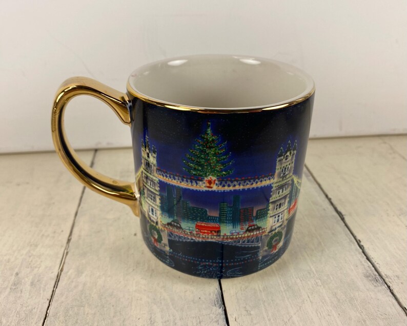 K&ouml;nnte beinhalten: Eine Keramik-Tasse mit goldenem Henkel und Rand. Die Tasse hat einen dunkelblauen Hintergrund mit einer Weihnachtsszene, darunter ein geschm&uuml;ckter Baum, Geb&auml;ude und eine Br&uuml;cke. Das Innere der Tasse ist wei&szlig;.