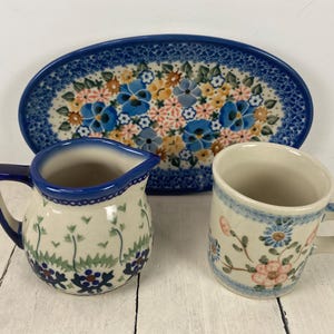 May include: A ceramic serving platter, creamer, and mug set. The oval platter and mug feature blue, yellow, and pink floral patterns. The creamer has a blue handle and rim, with a floral design.