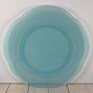 May include: A round, light blue glass plate with a scalloped edge. The plate has a smooth surface and a slightly translucent appearance. The plate is designed for serving food.