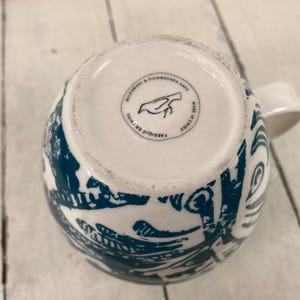 K&ouml;nnte beinhalten: Wei&szlig;e Keramiktasse mit tealblauem Muster und wei&szlig;em Henkel. Der Boden der Tasse tr&auml;gt die Aufschrift "Microwave & Dishwasher Safe" und "Made in China". Die Tasse hat ein Vogellogo.