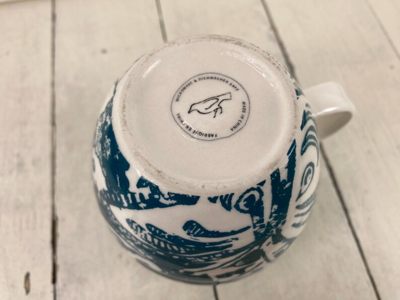 K&ouml;nnte beinhalten: Wei&szlig;e Keramiktasse mit tealblauem Muster und wei&szlig;em Henkel. Der Boden der Tasse tr&auml;gt die Aufschrift "Microwave & Dishwasher Safe" und "Made in China". Die Tasse hat ein Vogellogo.