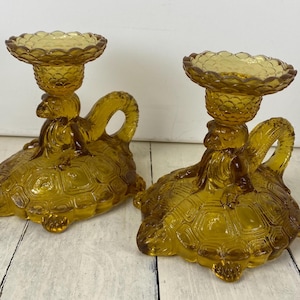 May include: Two amber-colored glass candle holders. Each candle holder is shaped like a turtle with a textured shell and a handle. The candle holders have a scalloped top to hold a candle. The candle holders are on a white surface.