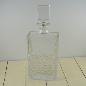 May include: A clear glass decanter with a square body and a square stopper. The decanter has a textured design on the bottom half.