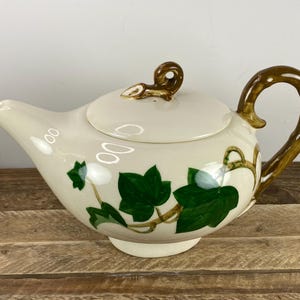 May include: Cream-colored ceramic teapot with a lid, handle, and spout. The teapot is decorated with green ivy leaves and brown vines. The handle and lid finial are brown. The teapot sits on a wooden surface.