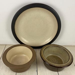 May include: A set of stoneware dishes, including a large plate with a black rim and two small bowls. The plate has a beige center, and the bowls are brown and olive green.