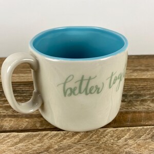 K&ouml;nnte beinhalten: Cremefarbene Keramiktasse mit hellblauem Innenraum und Henkel. Die Tasse tr&auml;gt den Schriftzug "better together" in hellgr&uuml;ner Schrift. Die Tasse steht auf einer Holzoberfl&auml;che.