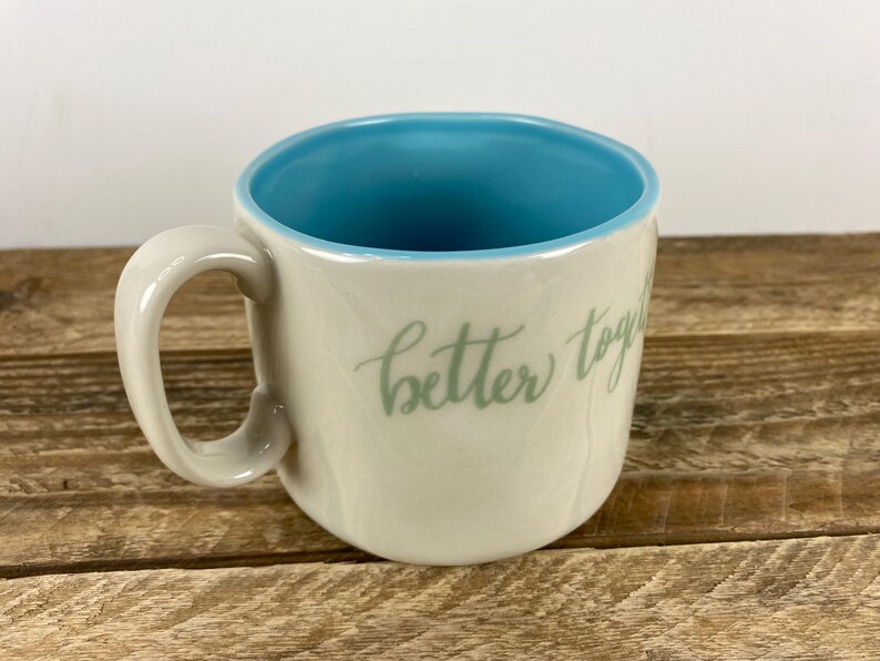 K&ouml;nnte beinhalten: Cremefarbene Keramiktasse mit hellblauem Innenraum und Henkel. Die Tasse tr&auml;gt den Schriftzug "better together" in hellgr&uuml;ner Schrift. Die Tasse steht auf einer Holzoberfl&auml;che.