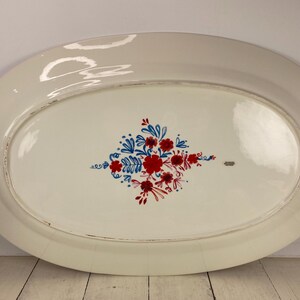 Op de afbeelding: Een ovale, witte keramische schaal met een bloemenontwerp. Het ontwerp toont rode en blauwe bloemen en bladeren in het midden. De schaal heeft een licht verweerde rand.