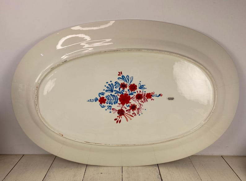 Op de afbeelding: Een ovale, witte keramische schaal met een bloemenontwerp. Het ontwerp toont rode en blauwe bloemen en bladeren in het midden. De schaal heeft een licht verweerde rand.