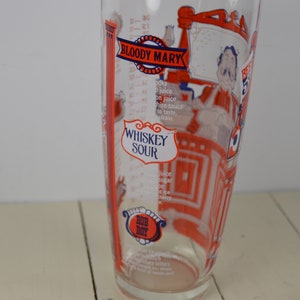 May include: A clear glass cocktail shaker with red and blue illustrations of a bartender and cocktail recipes for a Bloody Mary and Whiskey Sour. The shaker has measurements in ounces.