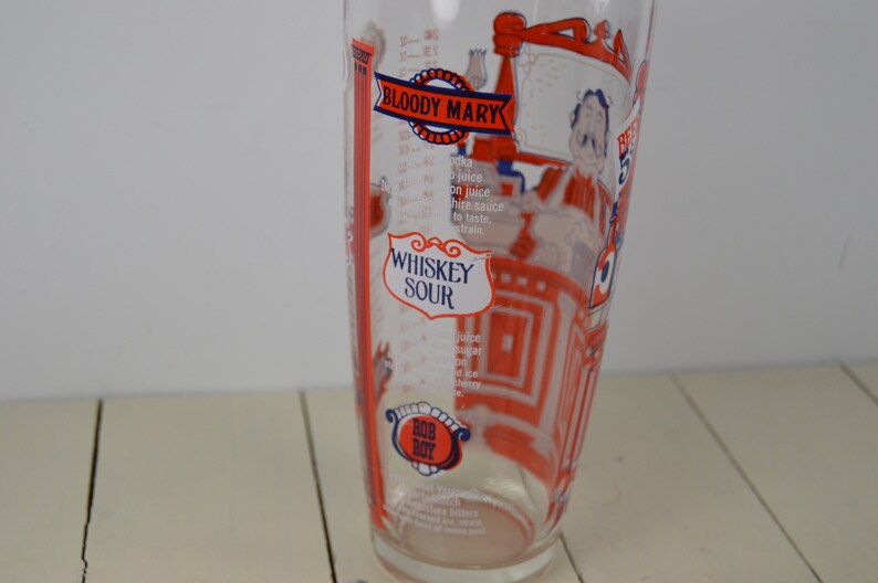May include: A clear glass cocktail shaker with red and blue illustrations of a bartender and cocktail recipes for a Bloody Mary and Whiskey Sour. The shaker has measurements in ounces.