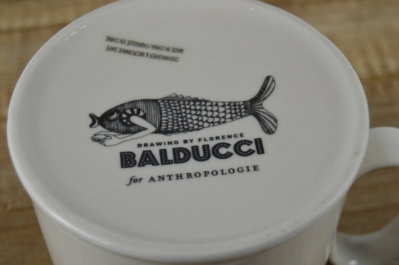 Pode incluir: Caneca de cer&acirc;mica branca com uma ilustra&ccedil;&atilde;o em preto e branco de um peixe com o texto "Drawing by Florence Balducci for Anthropologie" impresso na frente.