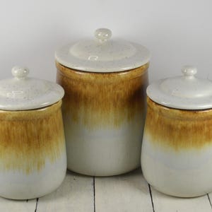 May include: Set of three ceramic canisters with lids. The canisters are white with a brown glaze that fades to white. The lids have a speckled white glaze.
