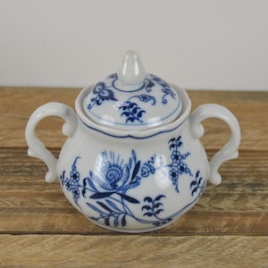 May include: A white porcelain sugar bowl with a blue and white floral pattern. The bowl has a lid and two handles.