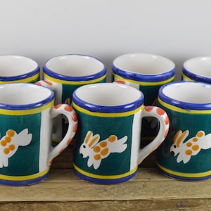 May include: Set of eight ceramic mugs with a green background and a white bunny with orange spots design. The mugs have a blue and yellow trim.