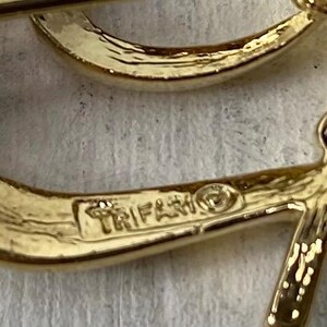 Op de afbeelding: Close-up van een goudkleurige broche met het woord "TRIFARI" erop gestempeld. De broche heeft een gebogen ontwerp met een getextureerd oppervlak. De broche staat op een lichtgeleurde achtergrond.