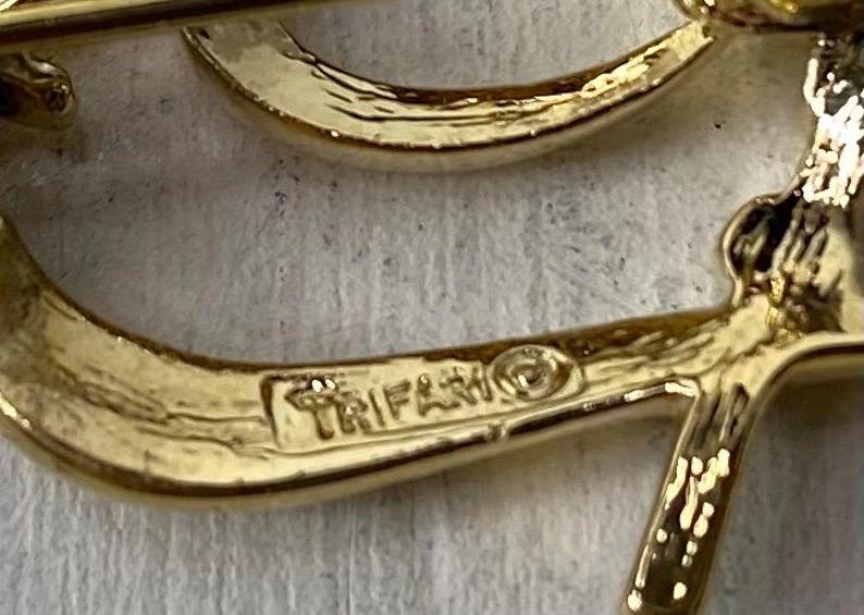 Op de afbeelding: Close-up van een goudkleurige broche met het woord "TRIFARI" erop gestempeld. De broche heeft een gebogen ontwerp met een getextureerd oppervlak. De broche staat op een lichtgeleurde achtergrond.