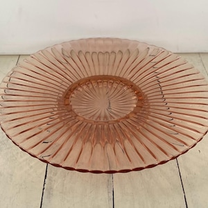 May include: A vintage pink glass serving plate with a ribbed, sunburst design. The plate has a raised center and a wide, flat rim. The color is a soft, muted pink, and the glass appears to be in good condition.