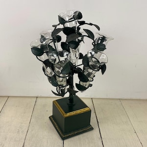 May include: A decorative centerpiece featuring multiple clear glass bud vases arranged amidst dark green metal leaves and stems. The vases are supported by a dark green base with gold trim, creating a unique floral display.